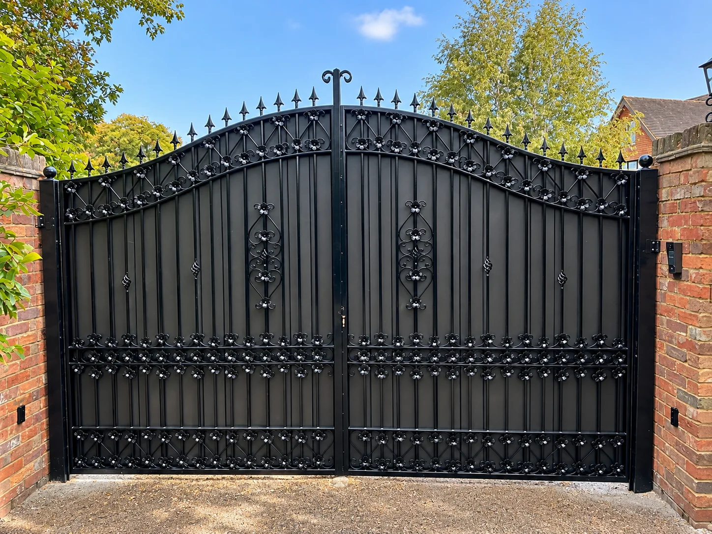 Home 47 Black arched panelled driveway gates with floral scrollwork and spear finials between brick pillars.